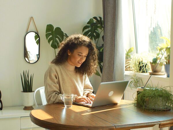 Frau mit Laptop an einem Holztisch. Im Hintergrund grüne Pflanzen.