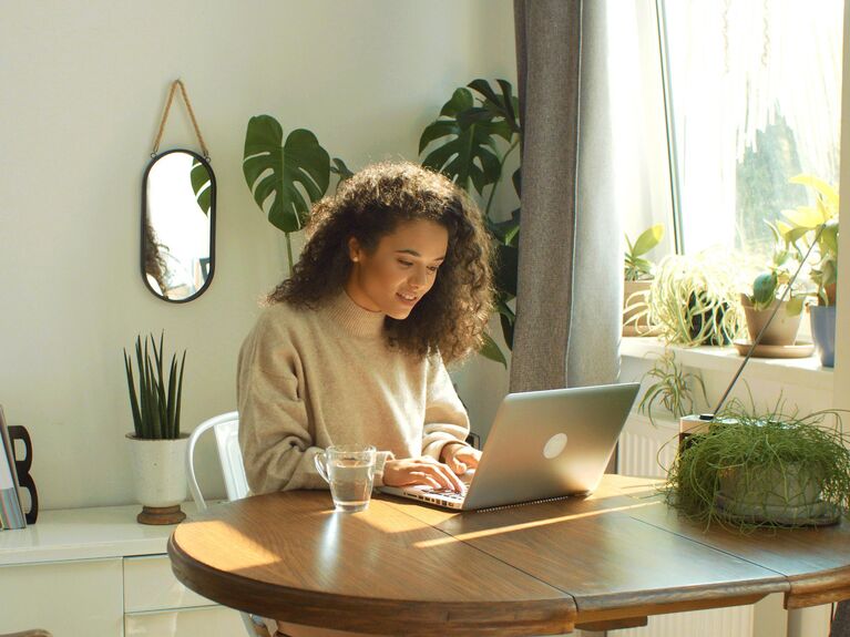 Frau mit Laptop an einem Holztisch. Im Hintergrund grüne Pflanzen.