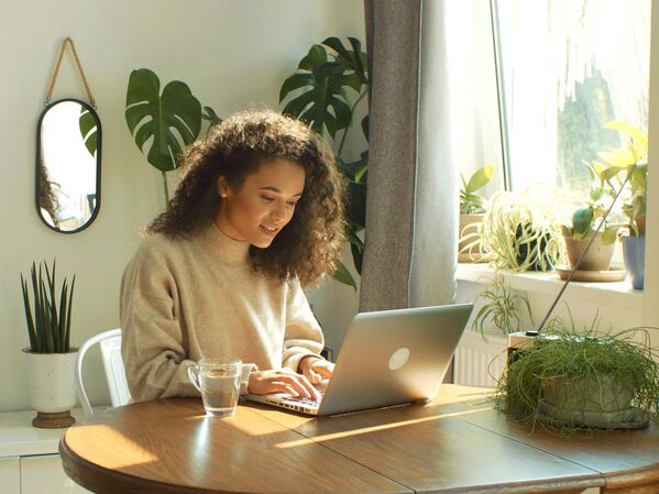 Frau mit Laptop an einem Holztisch. Im Hintergrund grüne Pflanzen.