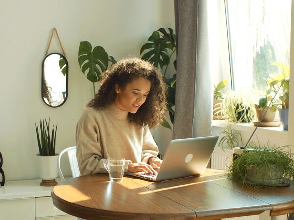 Frau mit Laptop an einem Holztisch. Im Hintergrund grüne Pflanzen.