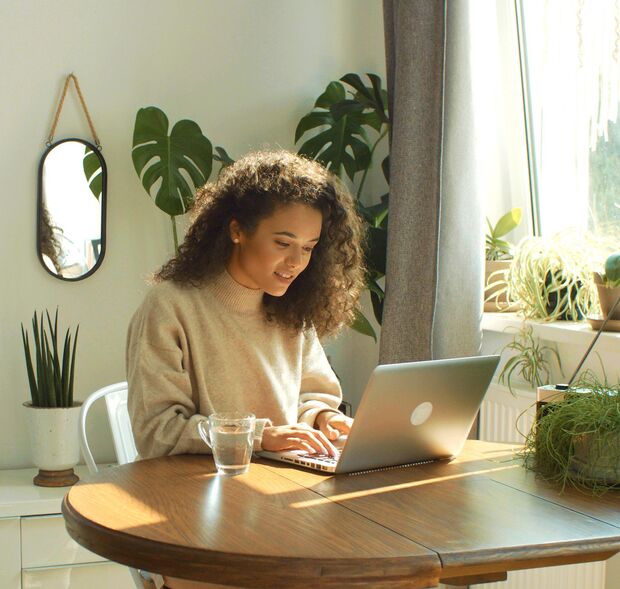 Frau mit Laptop an einem Holztisch. Im Hintergrund grüne Pflanzen.