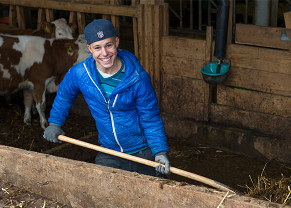 Junge Person mit blauer Jacke und Mütze arbeitet in einem Stall.
