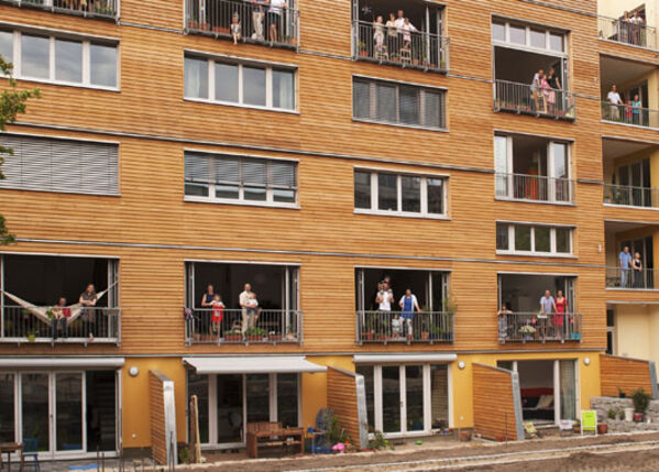 Modernes Haus mit Holzfassade und vielen großen Fenstern in denen Personen zu sehen sind.