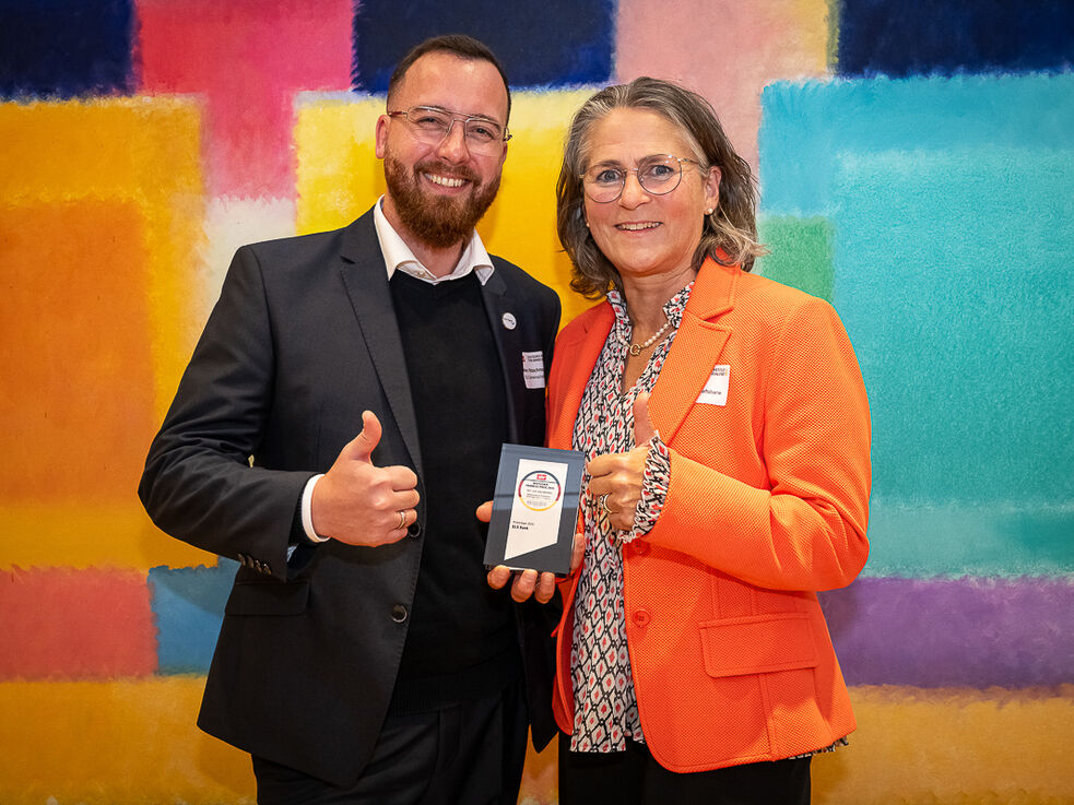 Zwei Menschen stehen lächelnd und mit erhobenem Daumen vor einer bunten Wand und halten eine Auszeichnung in der Hand.