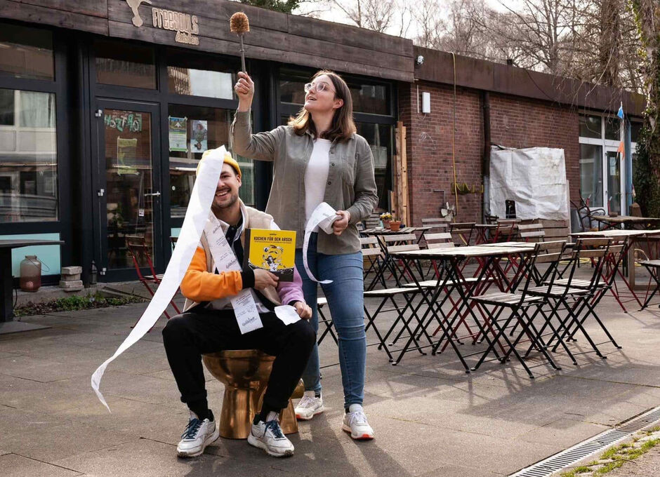 Zwei Personen mit Toilettenpapier und einer Toilettenbürste sitzen und stehen an den Tischen der Cafés im Freien.