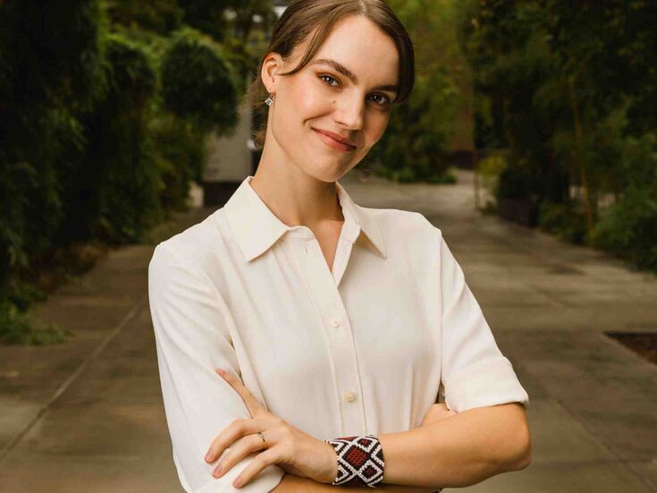 Eine weiblich gelesene Person in einem weißen Hemd steht im Freien mit verschränkten Armen und lächelt.