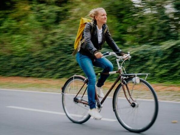 Weiblich gelesene Person fährt mit einem Fahrrad auf einer Straße vor begrüntem Hintergrund