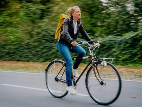 Weiblich gelesene Person fährt mit einem Fahrrad auf einer Straße vor begrüntem Hintergrund