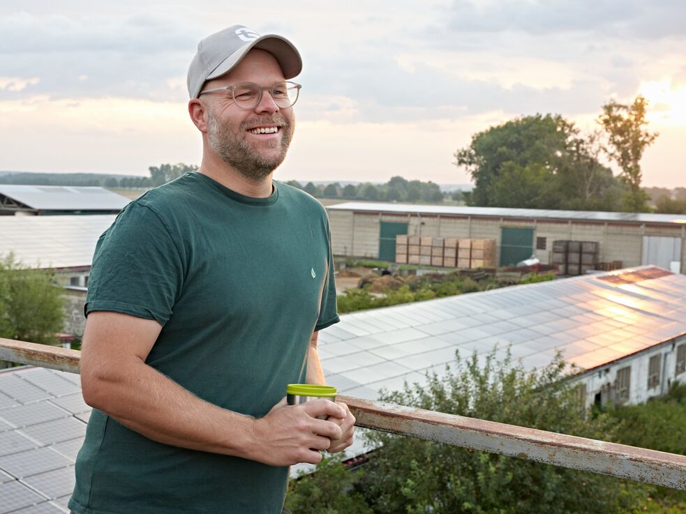 Mann mit Brille und Mütze, der einen Becher hält, steht bei Sonnenuntergang vor Sonnenkollektoren.