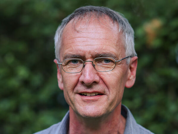 Älterer Mann mit kurzen grauen Haaren und Brille, der draußen vor einer Grünanlage steht.