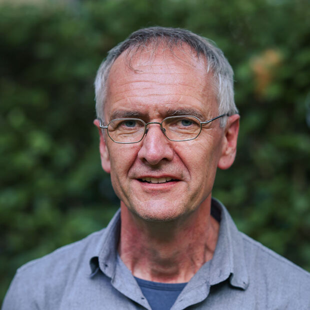 Älterer Mann mit kurzen grauen Haaren und Brille, der draußen vor einer Grünanlage steht.