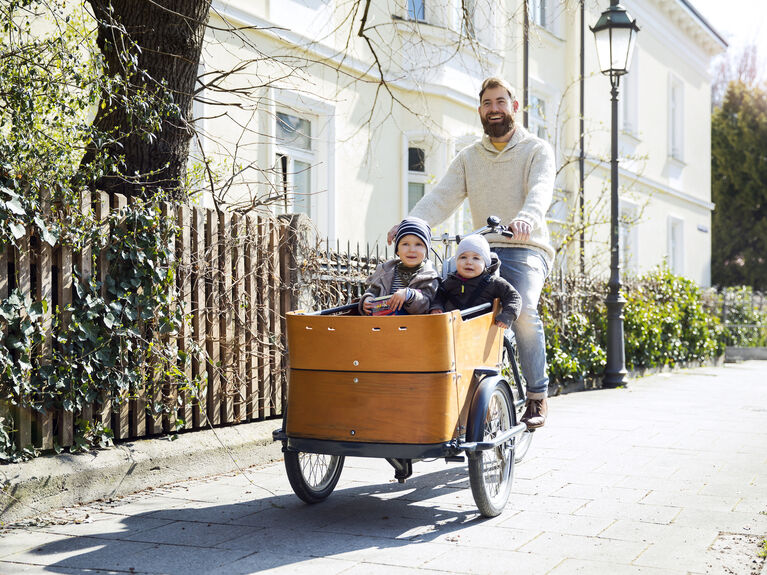 Ein Mann fährt auf einem Lastenfahrrad mit zwei Kindern, die in der vorderen Box sitzen, auf einem Bürgersteig.
