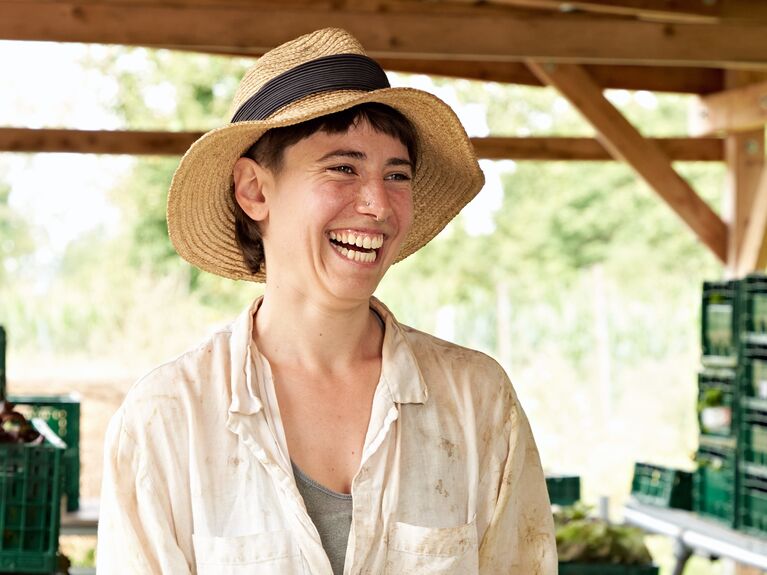 Person mit einem Strohhut, die lächelt und ein Produkt in der Hand hält, während sie mit einer anderen Person spricht.