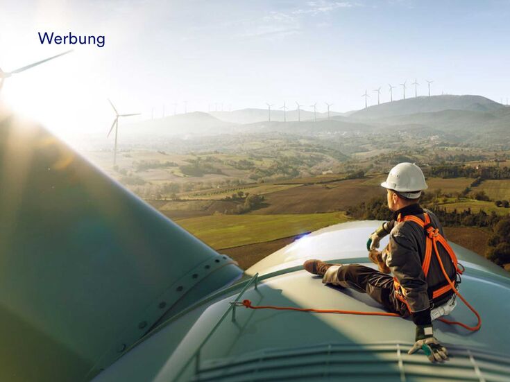 Eine männlich gelesen Person in Schutzkleidung sitzt bei Sonnenuntergang auf einer Windkraftanlage mit Blick auf einen Windpark.