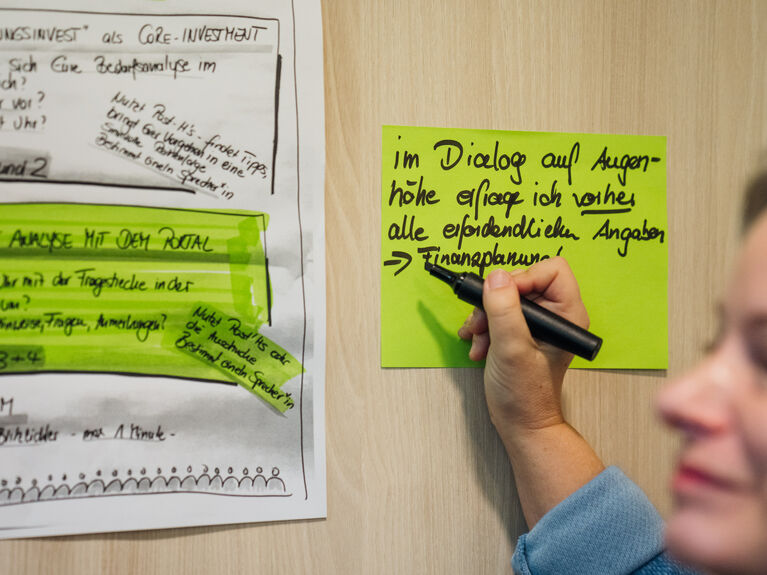 Eine Person schreibt auf einen grünen Zettel neben einer Tabelle mit hervorgehobenem Text an einer Wand.