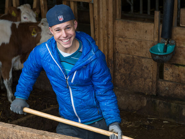 lachender Junge mit blauer Jacke und Mistgabel im Stall.