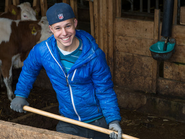 lachender Junge mit blauer Jacke und Mistgabel im Stall.