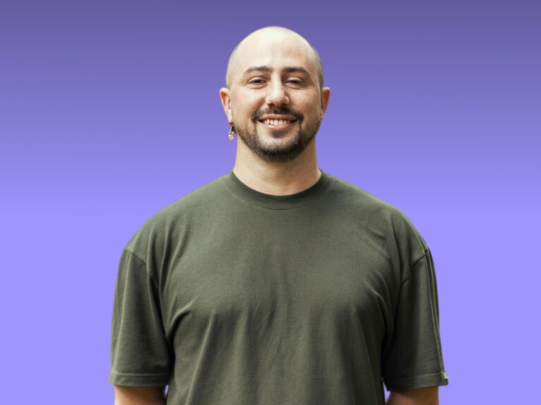 Eine Person in einem dunkelgrünen Hemd steht lächelnd vor einem lila Hintergrund mit Farbverlauf.