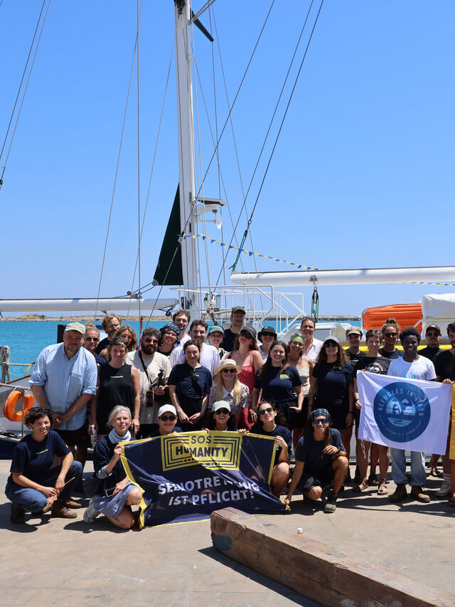 Eine große Gruppe steht auf einem Steg vor einem Segelboot und hält Transparente und Fahnen in den Händen.