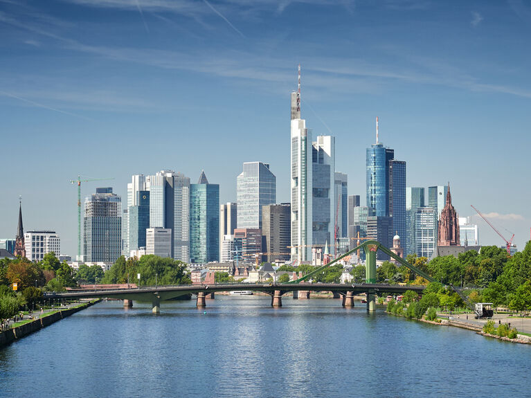Stadtpanorama mit modernen Wolkenkratzern und einem Fluss im Vordergrund unter blauem Himmel.