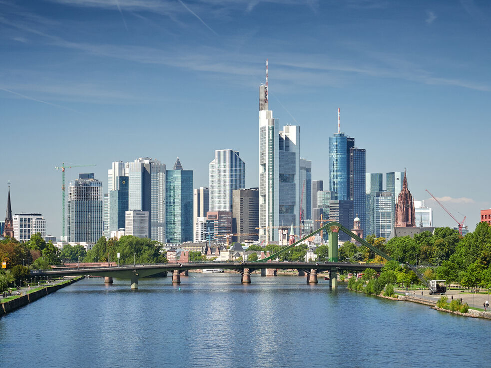 Stadtpanorama mit modernen Wolkenkratzern und einem Fluss im Vordergrund unter blauem Himmel.
