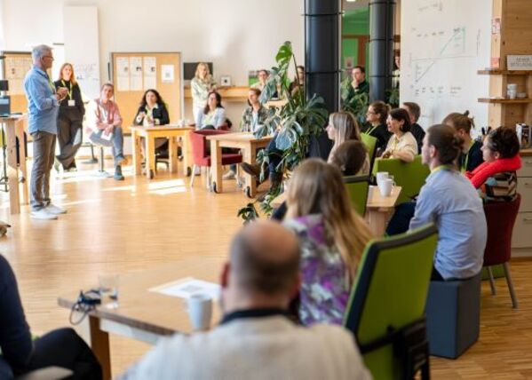 Eine Person links im Bild steht vor einer großen Gruppe an Menschen, die alle interessiert zuhören.