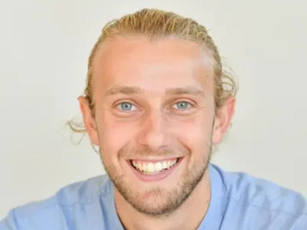 Ein Mann mit blondem Haar und Bart lächelt in die Kamera, er trägt ein hellblaues Hemd.