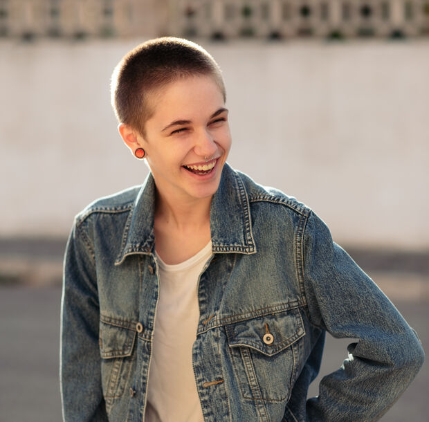 Person mit kurzen Haaren und Jeansjacke, die im Freien im Sonnenlicht lächelt.