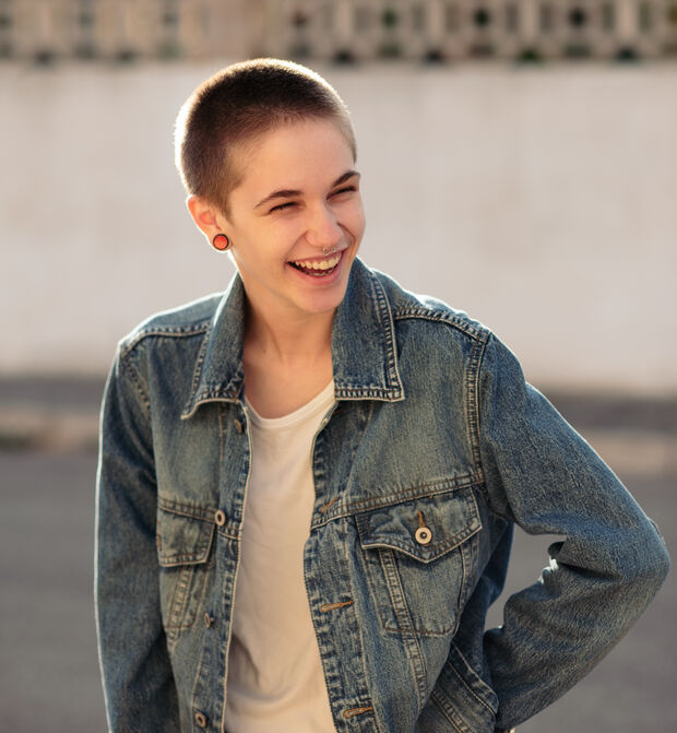 Person mit kurzen Haaren und Jeansjacke, die im Freien im Sonnenlicht lächelt.