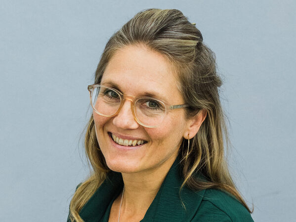 Frau mit Brille und grünem Hemd lächelt vor einem schlichten hellgrauen Hintergrund.