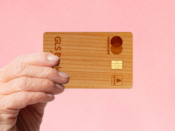 Hand einer älteren Person hält die Holzkarte der GLS Bank vor einen rosa Hintergrund