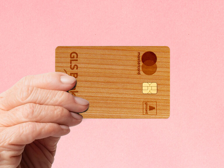 Hand einer älteren Person hält die Holzkarte der GLS Bank vor einen rosa Hintergrund