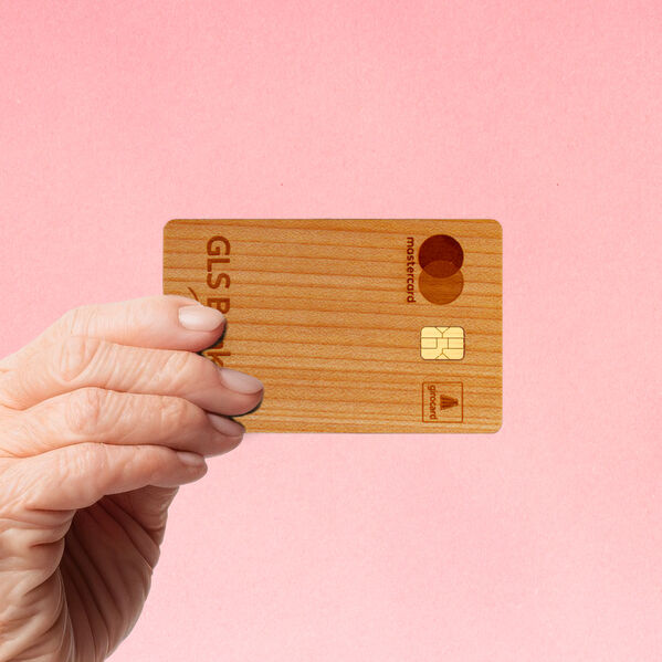 Hand einer älteren Person hält die Holzkarte der GLS Bank vor einen rosa Hintergrund