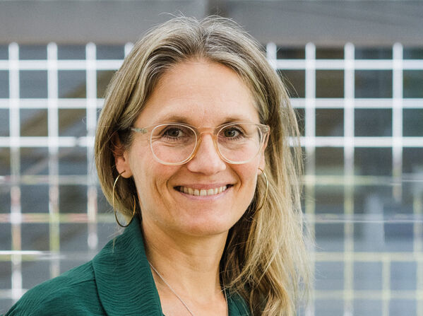 Eine Frau mit Brille und langen Haaren steht lächelnd vor einer Glaswand.