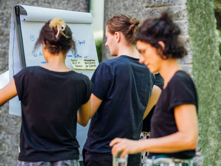Drei Personen arbeiten gemeinsam an einem Flipchart, auf dem mit blauem Marker Notizen geschrieben sind.