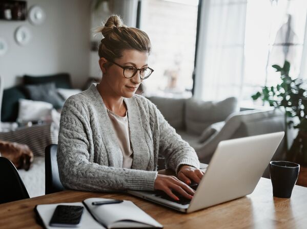 Frau arbeitet an einem Laptop an einem Holztisch mit einem Notebook und einem Telefon in der Nähe.