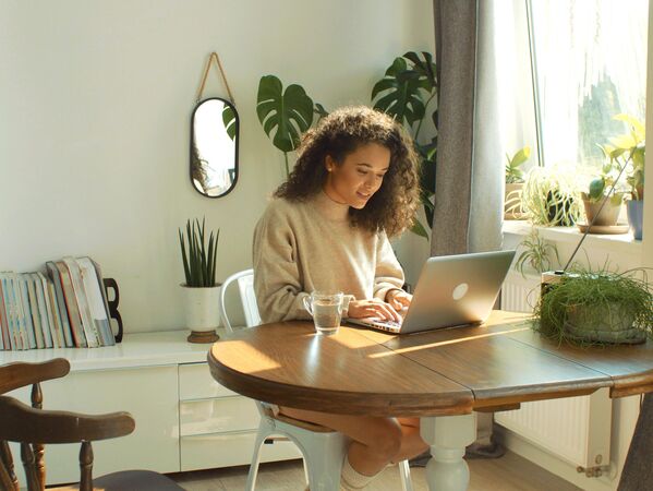 Frau mit Laptop an einem Holztisch. Im Hintergrund grüne Pflanzen.