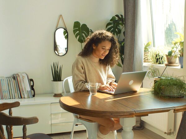 Frau mit Laptop an einem Holztisch. Im Hintergrund grüne Pflanzen.