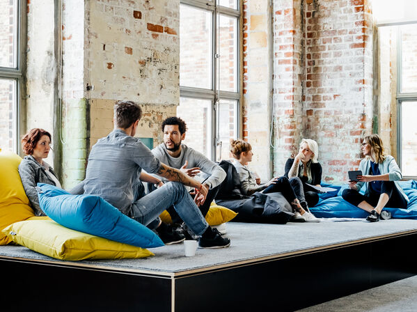 Sechs Personen sitzen und unterhalten sich auf Sitzreihen mit Sitzsäcken in einem modernen Büroraum.