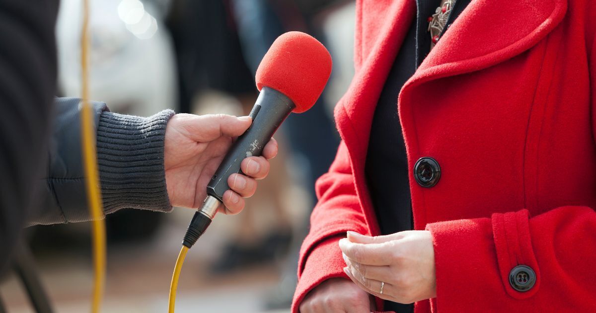 Einer Frau im roten Mantel wir ein Pressemikrofon hingehalten.