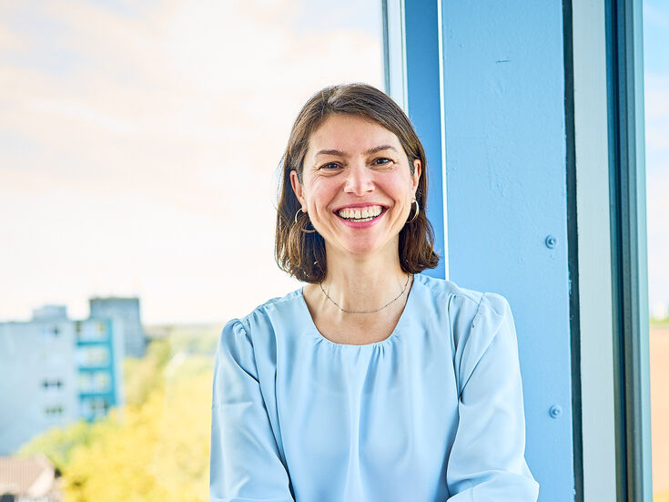 Eine Frau in hellblauer Bluse und roter Hose sitzt an einem Fenster und lächelt in die Kamera.