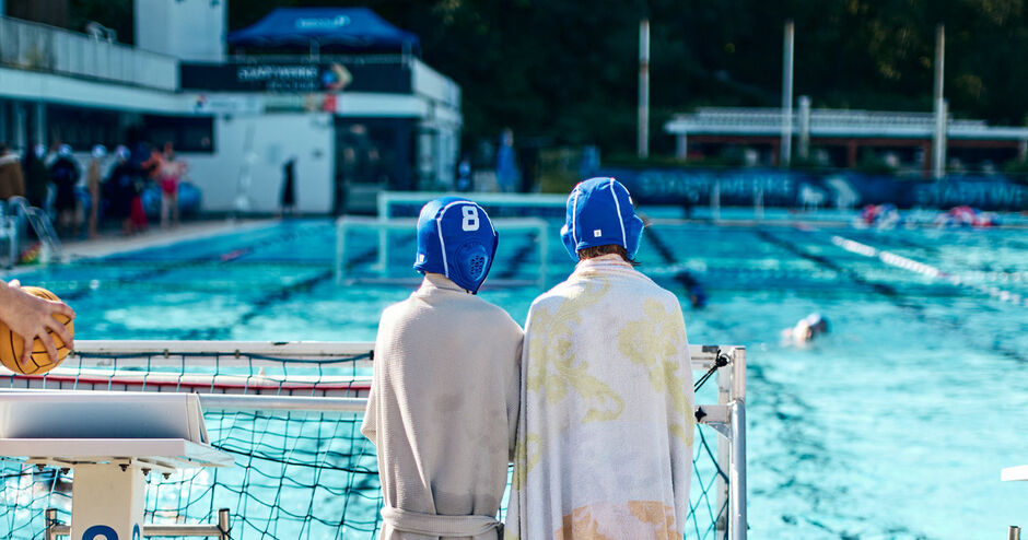 Zwei Wasserballspieler mit blauen Mützen und Handtüchern stehen an einem Außenbecken.