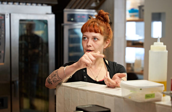 Eine Frau mit roten Haaren gestikuliert während eines Gesprächs in einer Großküche.