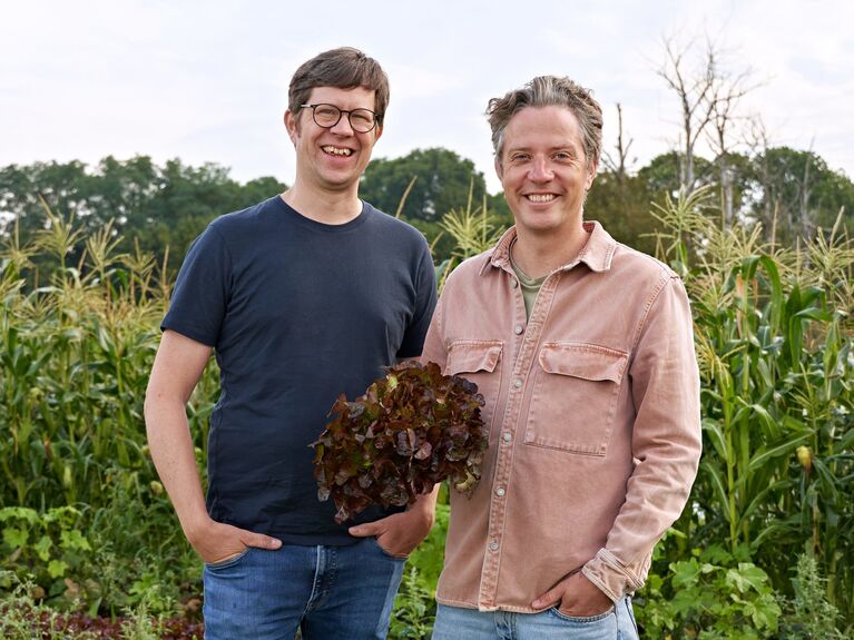 Zwei Personen stehen in einem Gemüsegarten und halten frisch geerntetes Blattgemüse in der Hand.