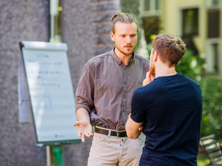 Zwei Männer führen ein ernstes Gespräch im Freien neben einem Flipchart mit Notizen.