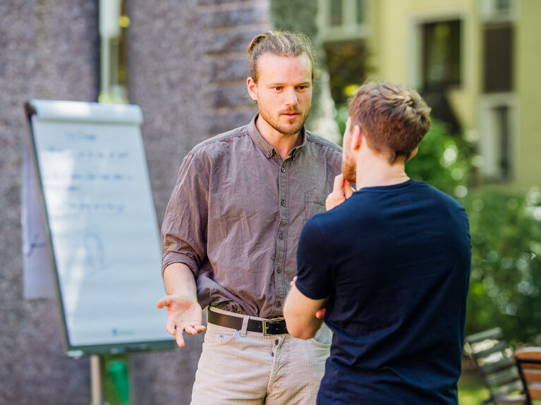Zwei Männer führen ein ernstes Gespräch im Freien neben einem Flipchart mit Notizen.