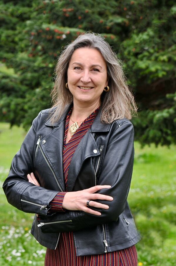 Frau in schwarzer Lederjacke steht im Freien mit verschränkten Armen und lächelt.