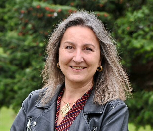 Frau in schwarzer Lederjacke steht im Freien mit verschränkten Armen und lächelt.