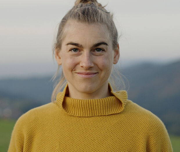 Eine Frau in einem gelben Pullover steht draußen, im Hintergrund sind Hügel zu sehen.