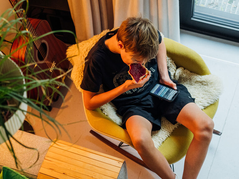 Ein Teenager sitzt auf einem Stuhl, isst und schaut auf ein Tablet in einem gemütlichen Wohnzimmer.